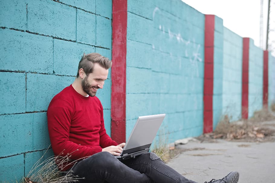 young, bearded, man, wearing, cherry, red, pullover, sitting, boundary wall, smilingly