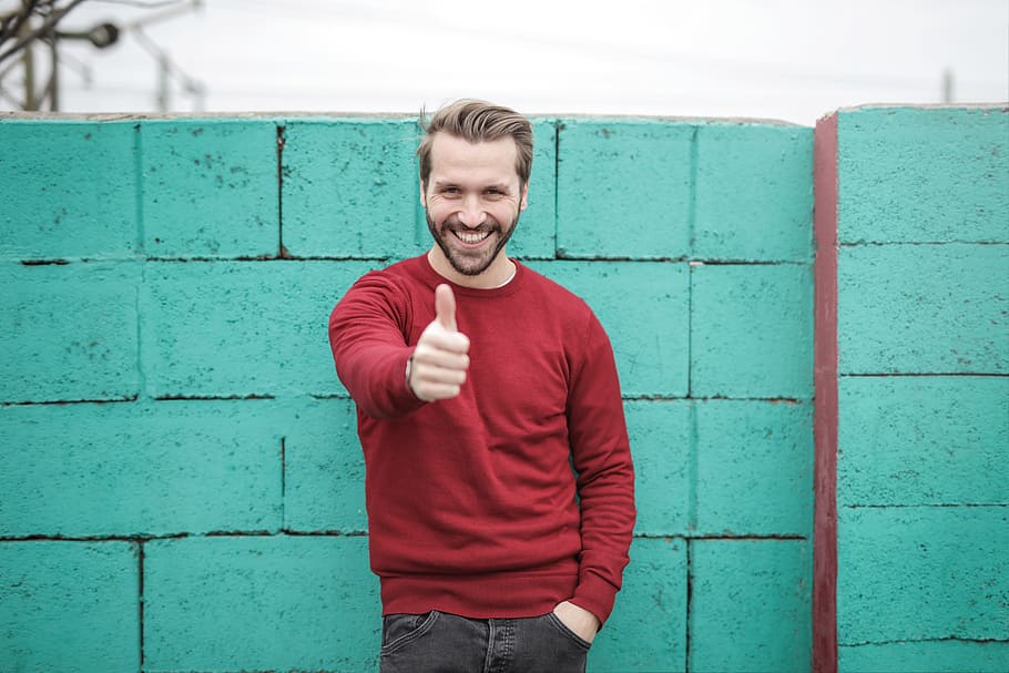 man, thumbs up, happy, smile, red, jumper, jersey, blue, brick, wall.jeans