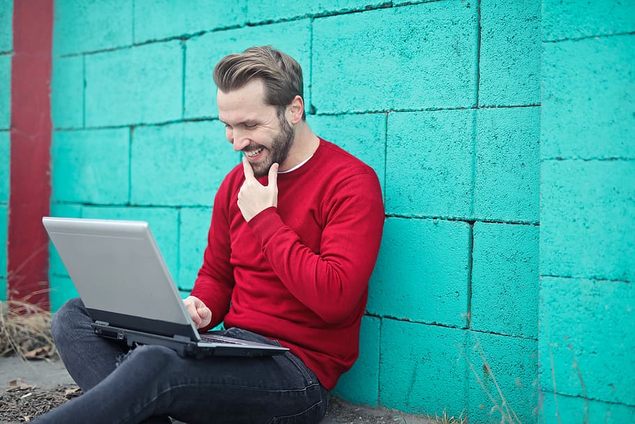 adult man, red, sweatersmiling, using, laptop, turquoise, brick wall, 30-35 years old, Adult, Outdoors